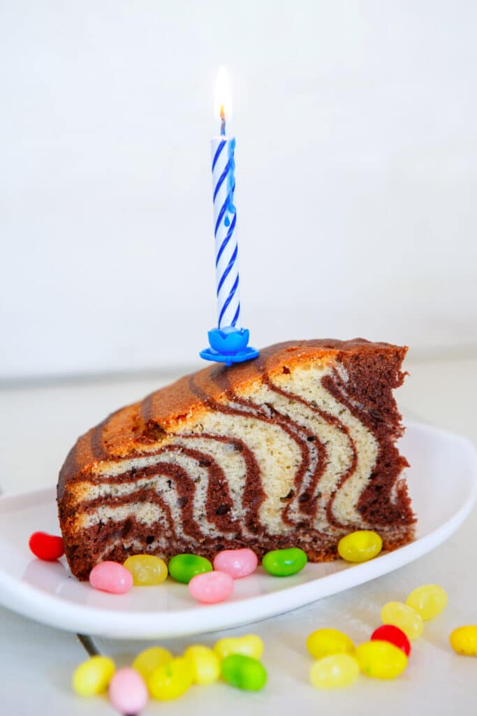 Tranche de gâteau marbré avec bougie d'anniversaire allumée, entourée de bonbons colorés sur une assiette blanche.