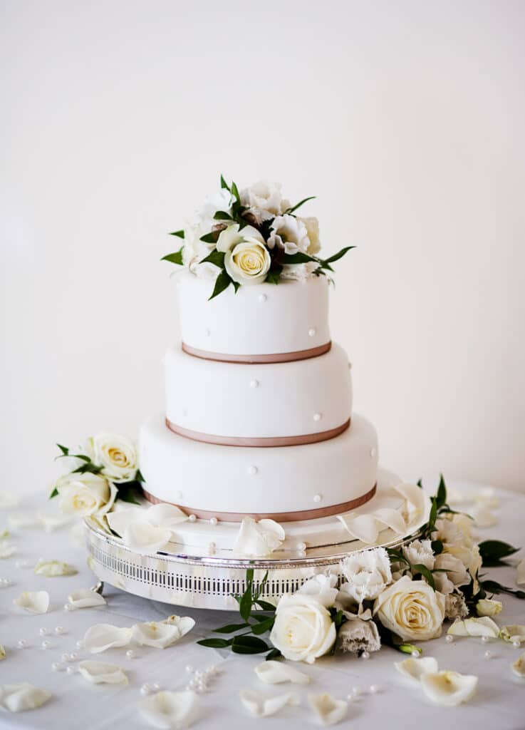 Gâteau de mariage blanc à trois étages décoré de roses, perles et rubans bruns sur un support argenté, avec pétales.