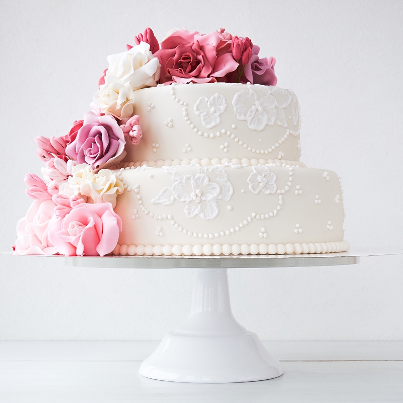Élégant gâteau de mariage blanc à deux étages, orné de roses et fleurs de sucre roses et blanches, posé sur un support blanc.