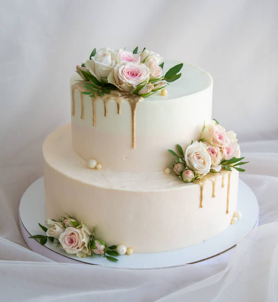 Gâteau deux étages crème et blanc, décoré de roses pastel, de coulures dorées brillantes et de perles, sur un voile clair.