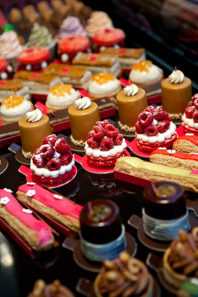 Un étalage coloré de pâtisseries françaises. On y voit des tartelettes aux framboises, des gâteaux au caramel, des éclairs roses et jaunes, et des cupcakes variés.