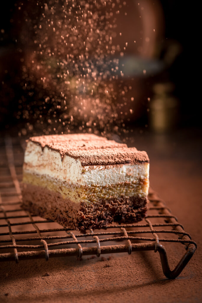 Part de tiramisu ou dessert chocolaté multicouche sur grille, généreusement saupoudré de cacao, créant une atmosphère gourmande.