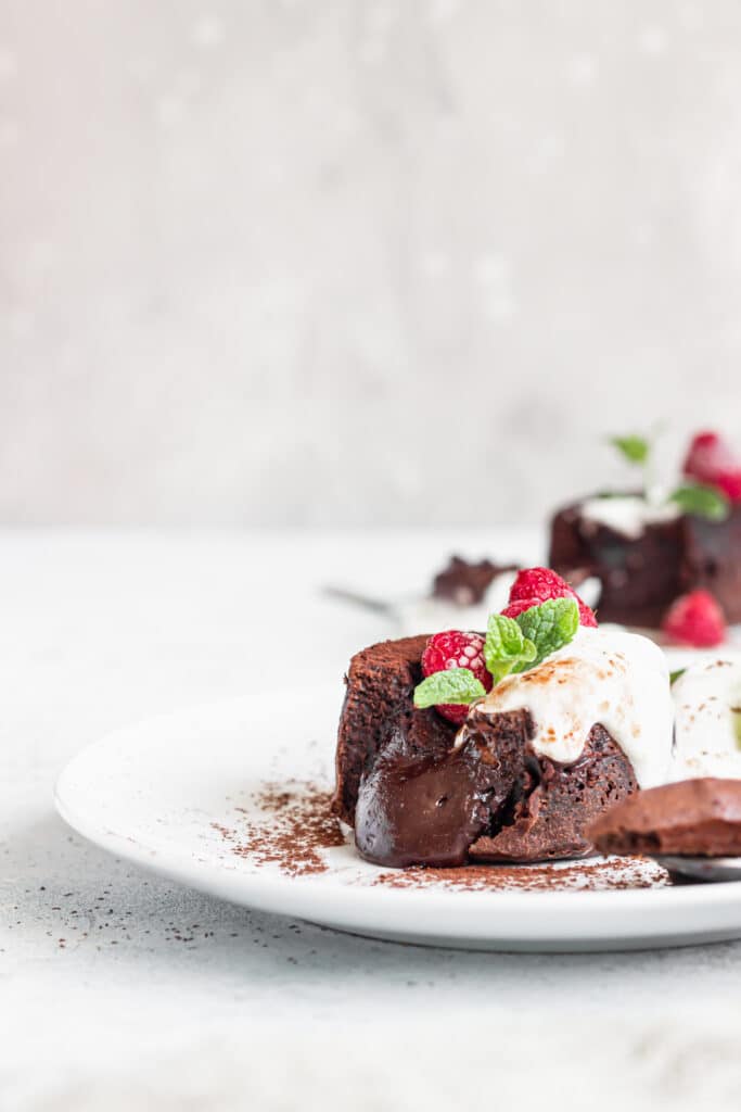 Fondant au chocolat coulant, framboises, crème et feuille de menthe sur assiette blanche. Coulis onctueux, poudre de cacao.
