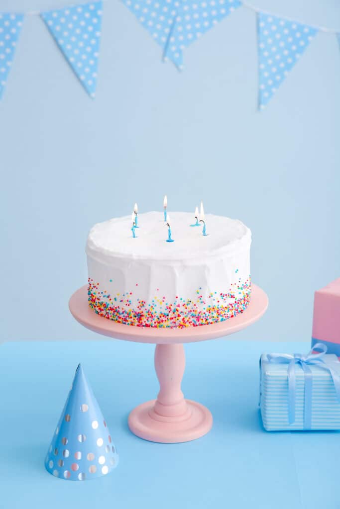 Gâteau d'anniversaire blanc avec vermicelles colorés et cinq bougies bleues allumées. Table bleue, cadeaux emballés, chapeau de fête et guirlande en arrière-plan.